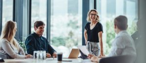A group of business people sit around a conference table with one standing to lead a funny topic