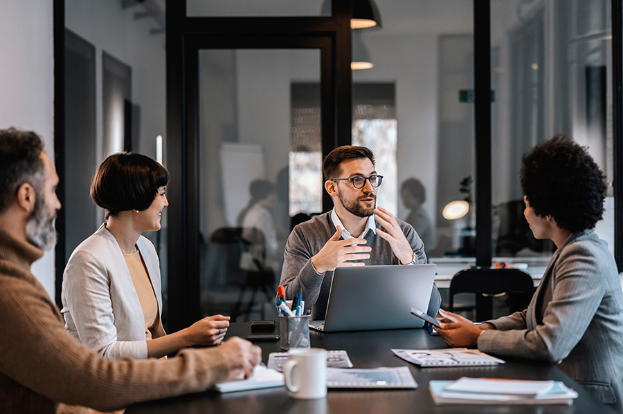 Four professionals sit at the end of a long conference table looking at the main speaker