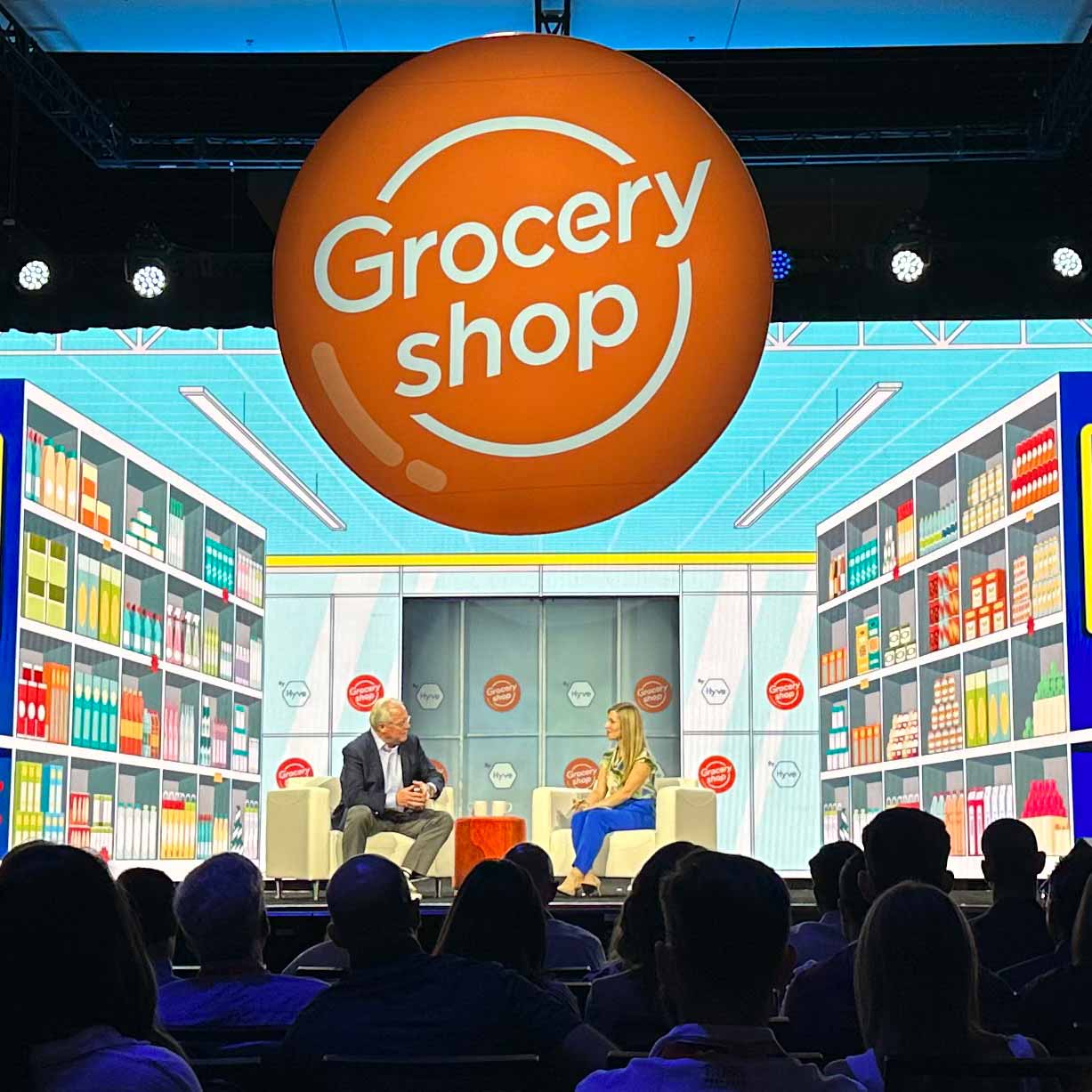 Two well dressed executives in suits sit in front of a LED display of a grocery store.