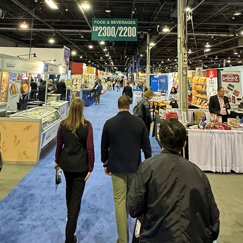 three people in dark jackets walk on blue carpet in a tradeshow isle at the PLMA 2023 tradeshow