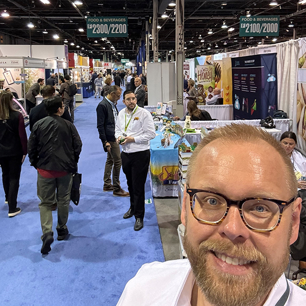 Portrait of blonde gentleman with a beard and white shirt in a tradeshow isle at the PLMA 2023 tradeshow