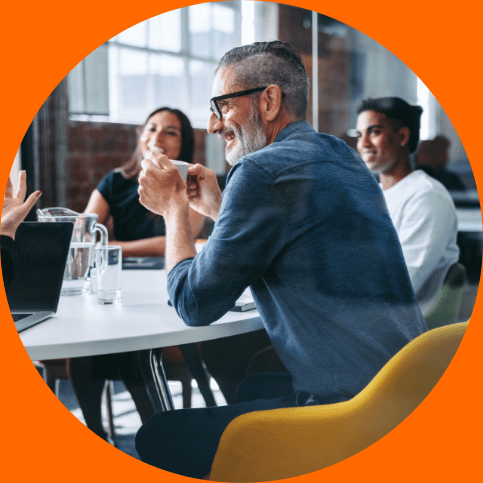 Three business people having a discussion, the foreground includes a gray haired man wearing a blue shirt laughing