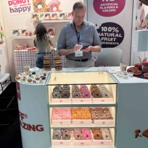 gentleman wearing a check shirt checks his phone in front of a case of brightly colored donuts.
