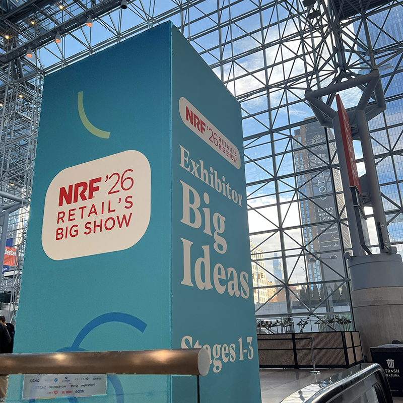 header wIMG_6484 Large teal event signage displaying "NRF '26 Retail's Big Show" with "Exhibitor Big Ideas" and "Stages 1-3" text at the Javits Center entrance, glass ceiling architecture visible above.
