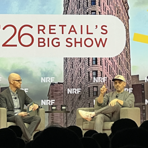 Two executives in fireside chat on NRF main stage, one bald with glasses in gray suit, one in casual sweater and cap gesturing while speaking, NYC skyline backdrop behind them.