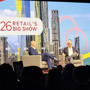 Executive interview on NRF '26 main stage featuring two men in suits, colorful New York City skyline graphics displayed on LED screens behind the conversation.