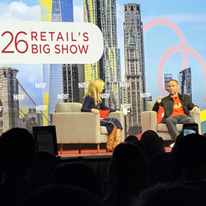Female interviewer speaking with male executive wearing orange shirt on NRF stage, NYC building graphics and "Retail's Big Show" branding prominently displayed.