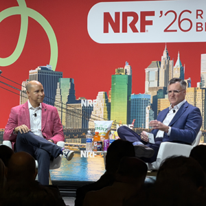 Two executives on NRF stage, one in bold pink blazer with white shirt, one in navy suit, Simply brand products visible on coffee table between them.