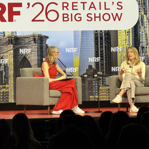 Wide shot of female executive interview on NRF stage, both women seated in armchairs, audience watching presentation about retail leadership.