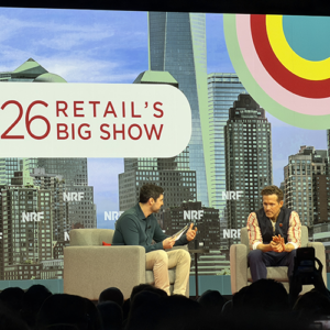 Two male executives in fireside chat at NRF '26, one in green polo with khakis holding notes, one in patterned jacket, colorful bullseye graphic and NYC skyline on screen behind them.