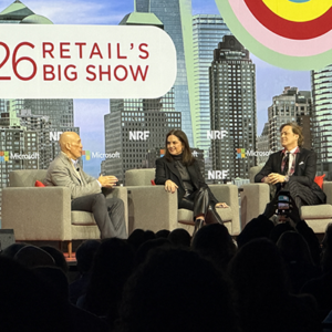 Three-person panel on NRF main stage with Microsoft sponsorship visible, bald executive in gray suit speaking while female panelist in black and male colleague listen, audience phones capturing the moment.