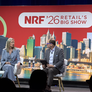 Executive interview on red-themed NRF stage, woman in light blue suit speaking with male moderator wearing purple-framed glasses and holding tablet, vibrant NYC skyline graphics behind them.