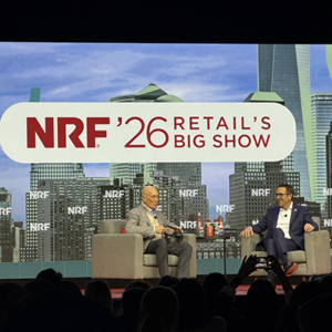 Two executives in conversation on NRF main stage, both in gray suits seated in armchairs, One World Trade Center prominently featured in backdrop graphics, audience silhouettes in foreground.