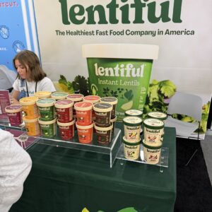 Tiered display of "lentiful" instant lentil cups beside a giant cup prop and a seated booth attendant.
