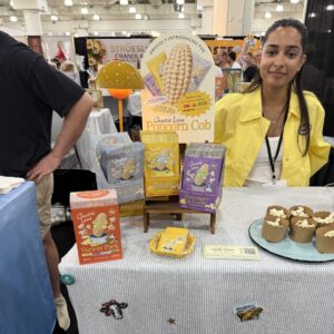 Woman in yellow smiles behind a table of "Cheerie Lane" popcorn products and dessert cups at a booth.