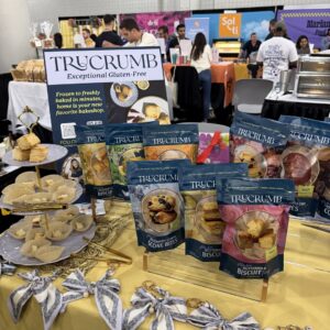 Row of "TRUCRUMB" gluten-free baked goods packages and a tiered tray of scones on a yellow tablecloth.