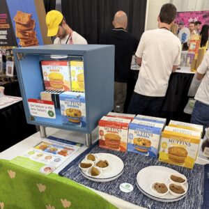 Booth table with a blue mini-freezer and "unrefined MUFFINS" boxes beside plates of muffin samples.