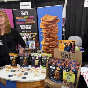 "every body eat" snack packages and sample cups on a table before banners reading "Crunch Without Compromise."
