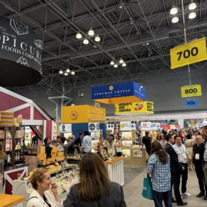 Busy trade show aisle with a cheese display booth, hanging 'World's Best Cheeses' and 'Schuman Cheese' signs.
