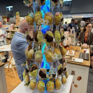 Tall tiered tower of decorated madeleine pastries with flowers and blue lighting on a booth display.