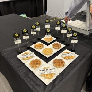 Waffles on white plates with "Le Waf." flavor signs and "Display Only - Please Do Not Eat" cards on a black table.