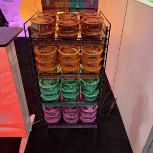 Black wire display rack stacked with colorful "Long Weekend" premium instant soup cups in orange, teal and purple.