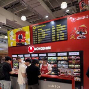 "Nongshim" booth styled as an airport gate with a departures board and shelves of ramen and snacks.