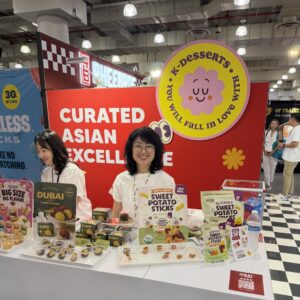 Two women at a booth with sweet potato sticks and Dubai cookie samples under a 'K-Desserts' and 'CURATED ASIAN EXCELLENCE' sign.