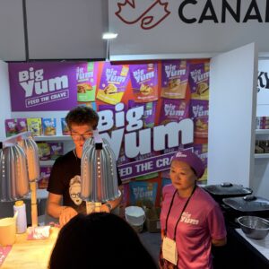 Two staff demoing food at the purple 'Big Yum' booth with product pouches on the back wall and a heat lamp on the counter.