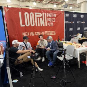 Four men seated in a filmed interview with cameras on tripods at the 'LOOPINI' protein-packed pizza booth.
