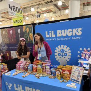 Two women behind a blue 'LiL BUCKS' booth table covered with buckwheat product boxes and tasting cups.