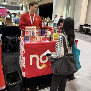 Exhibitor in a red blazer serving samples at a red 'noodo' booth as a visitor with a tote bag looks on.