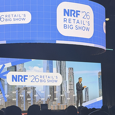 Wide shot of keynote presentation at NRF 2026 showing speaker on stage beneath massive curved screen displaying event branding, audience watching from below.