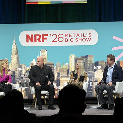 Four panelists on teal-themed NRF stage with Chrysler Building backdrop, female moderator engaging three retail executives including man in all black and woman with long blonde hair.